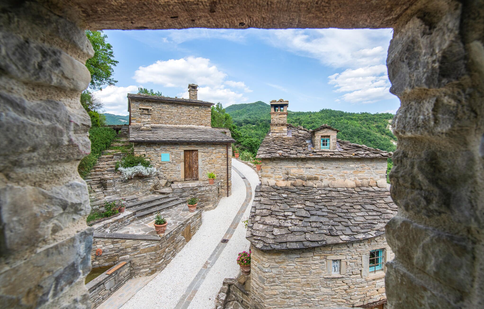 A stone pathway leading to traditional stone buildings with chimneys, surrounded by greenery and hills.