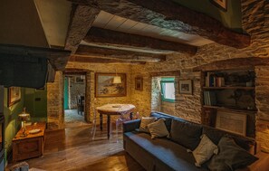 Ein gemütliches Zimmer mit Steinwand, Holzbalken, einem runden Tisch und einer Couch mit Kissen.