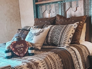 Ein Bett mit einem gemusterten Überwurf, mehreren Kissen, einer herzförmigen Schachtel und einer Flasche.