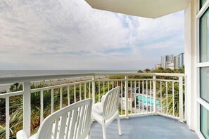 Ein Balkon mit weißen Stühlen, einem Pool und Blick auf Strand und Gebäude.