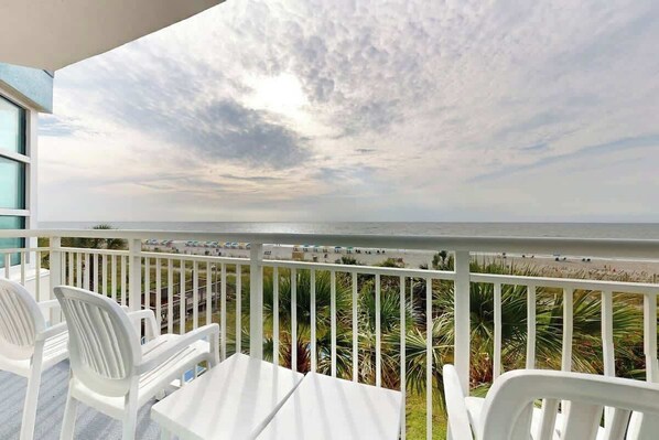 Un balcon avec des chaises blanches et une table, donnant sur une plage bordée de palmiers et offrant une vue sur l’océan.