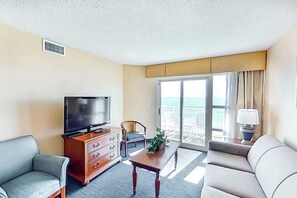 Quarto de hotel com televisão, sofá, poltrona, mesa e vista para o mar através da varanda.