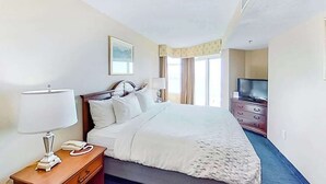 Ein Hotelzimmer mit einem Bett, einem Nachttisch mit Lampe, einem Telefon, einem Fernseher auf einer Kommode und einem Fenster mit Vorhängen.