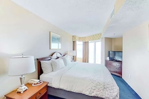 Quarto de hotel com cama, mesa de cabeceira com luminária, telefone, televisão em uma cômoda e uma janela com cortinas.