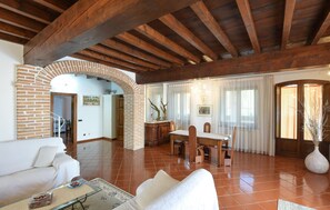 A spacious living area with a brick fireplace, wooden ceiling beams, a dining table with chairs, and a large window with sheer curtains. - Stunning home in Goito with WiFi (Goito)