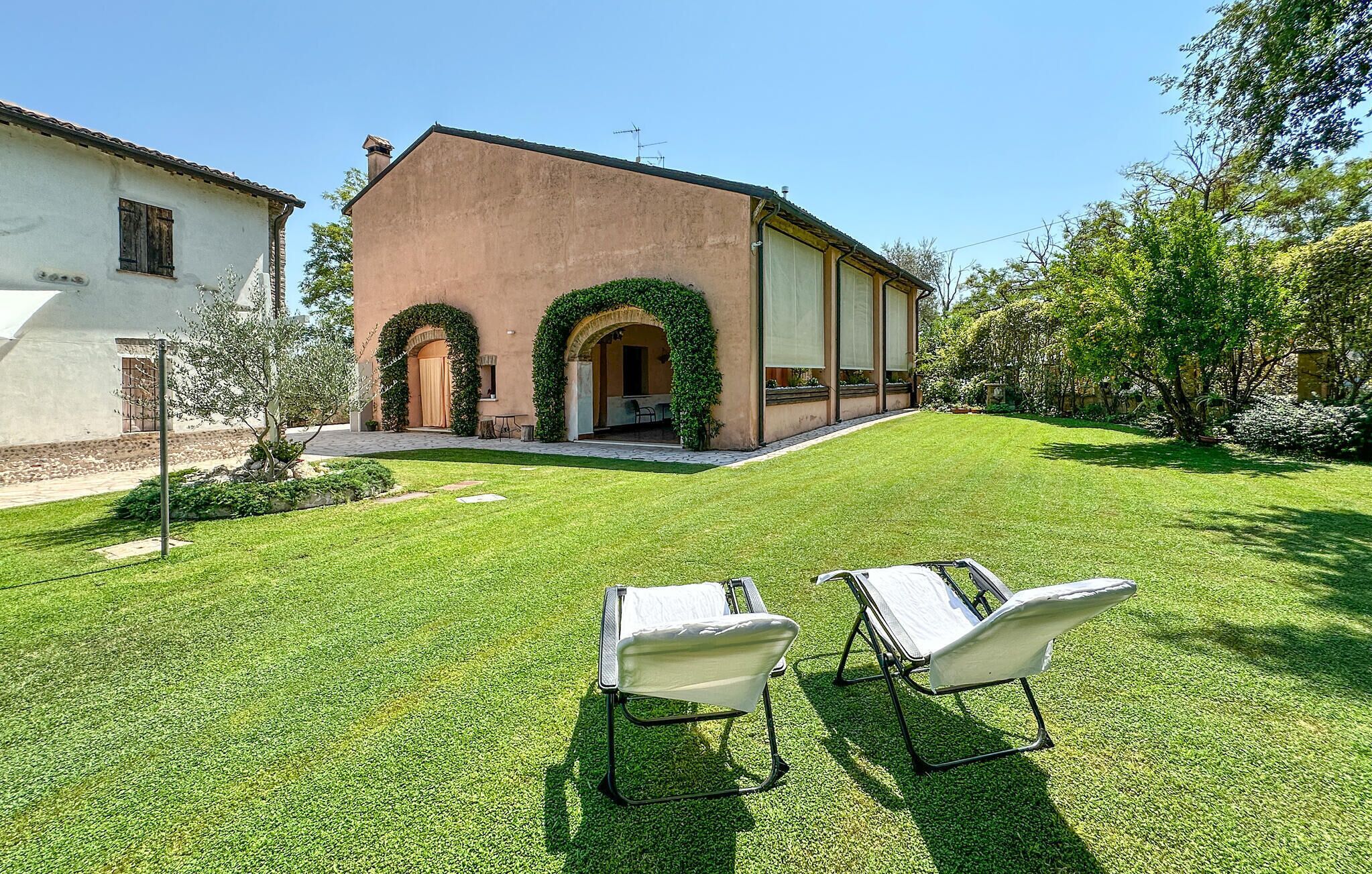 A building with two arched doorways, a covered patio area, and a well-maintained lawn with two white lawn chairs.