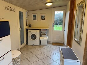 Een wasserette met een wasmachine, droger en een klein tafeltje met een kleedje. Er hangen foto's in lijsten aan de muur en een deur die naar buiten leidt.