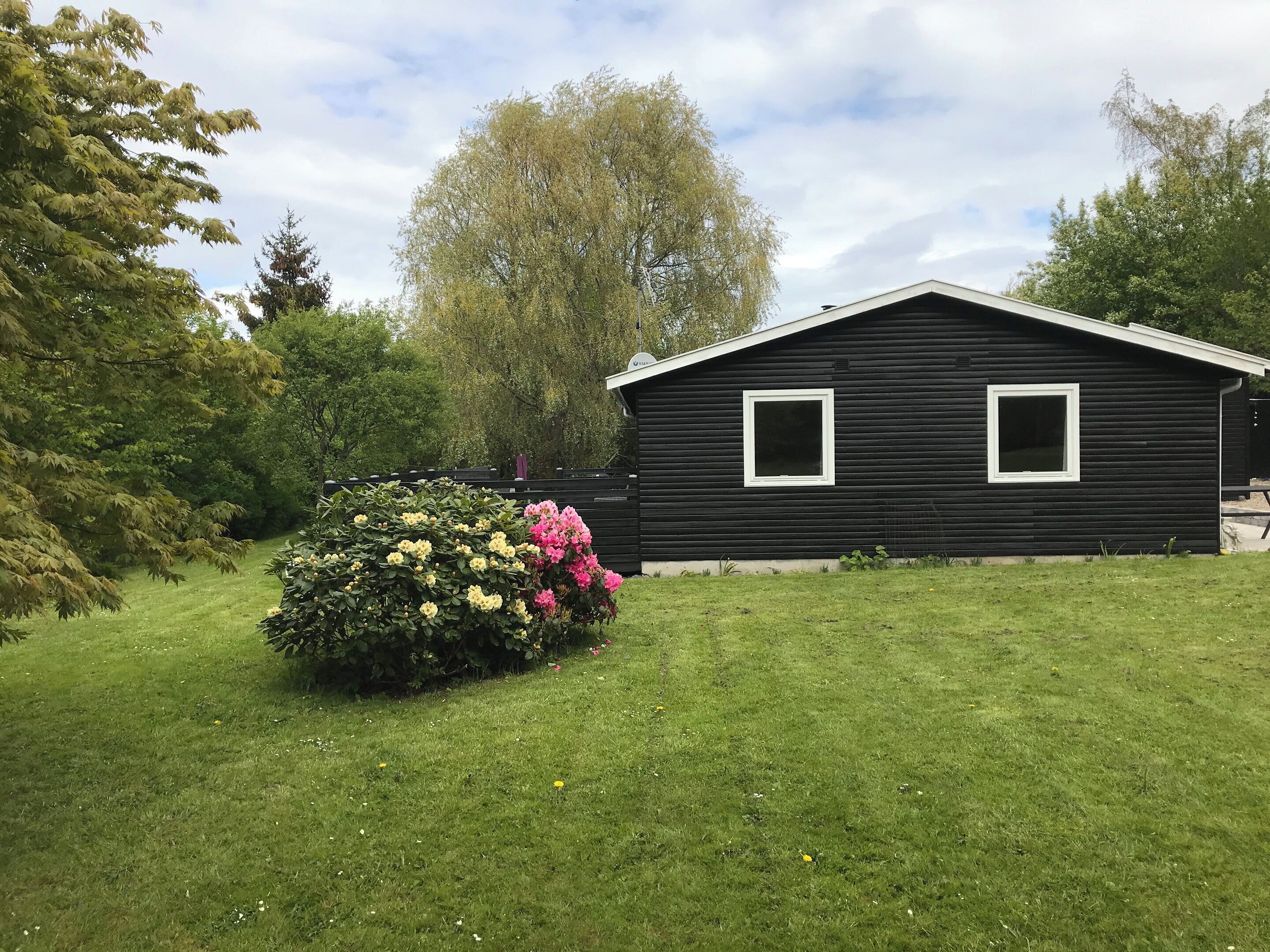 A black wooden house with a white window, surrounded by a well-maintained lawn and flowering bushes.