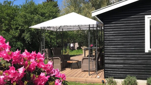 En træhytte med en overdækket terrasse, fletmøbler og en have med lyserøde blomster.