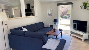 En moderne stue med en mørkeblå sofa, et træbord, et fjernsyn på et stativ og et stort vindue med udsigt til grønne områder.