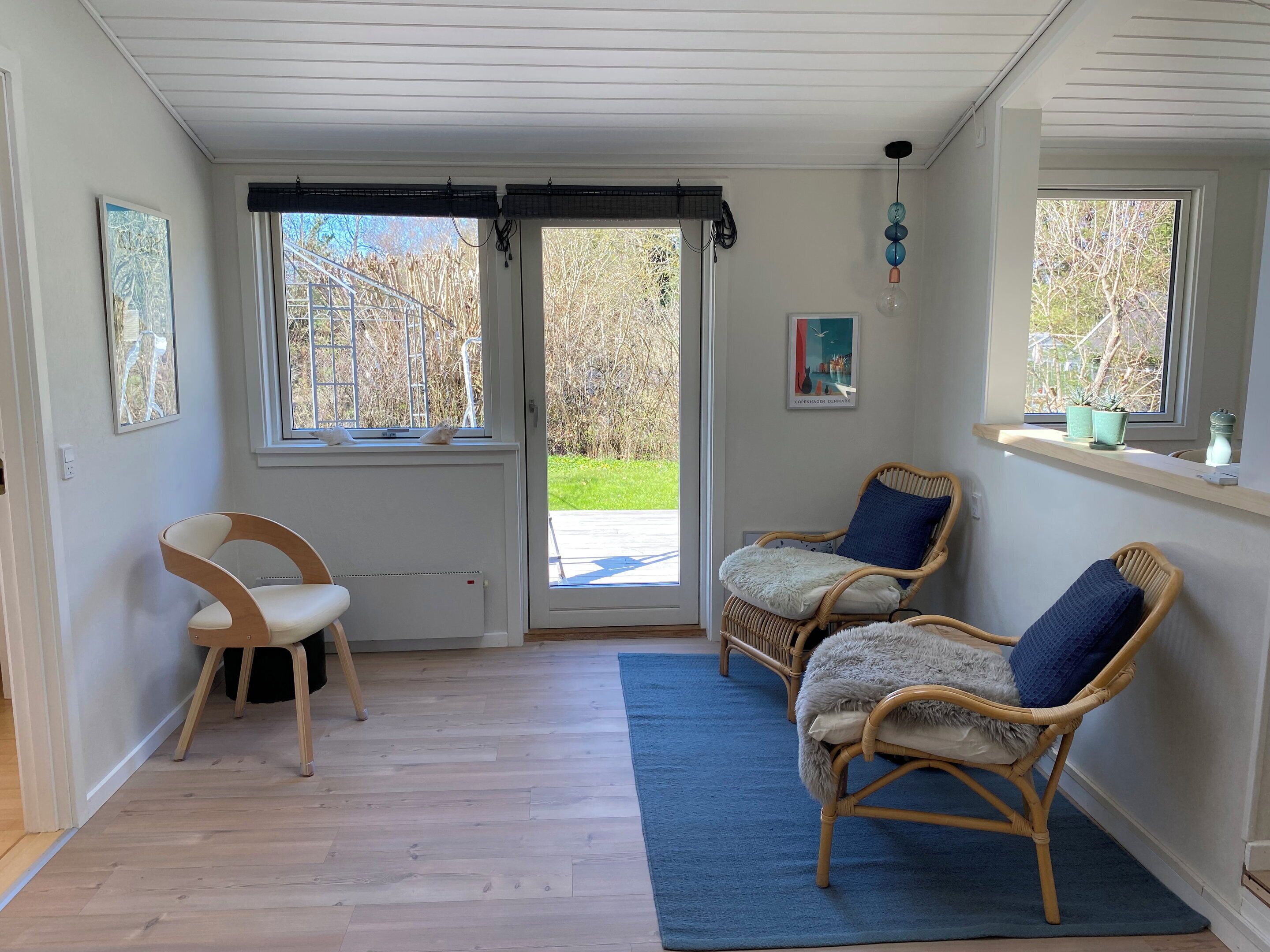 Um quarto com chão de madeira, duas cadeiras de vime com almofadas, uma cadeira branca e um tapete azul.
