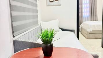 A hotel room with a bed, a small table with a plant, a window with blinds, and a door leading to another room.