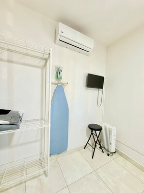 A room with a white air conditioner, a flat-screen TV mounted on the wall, a radiator, and a blue ironing board.