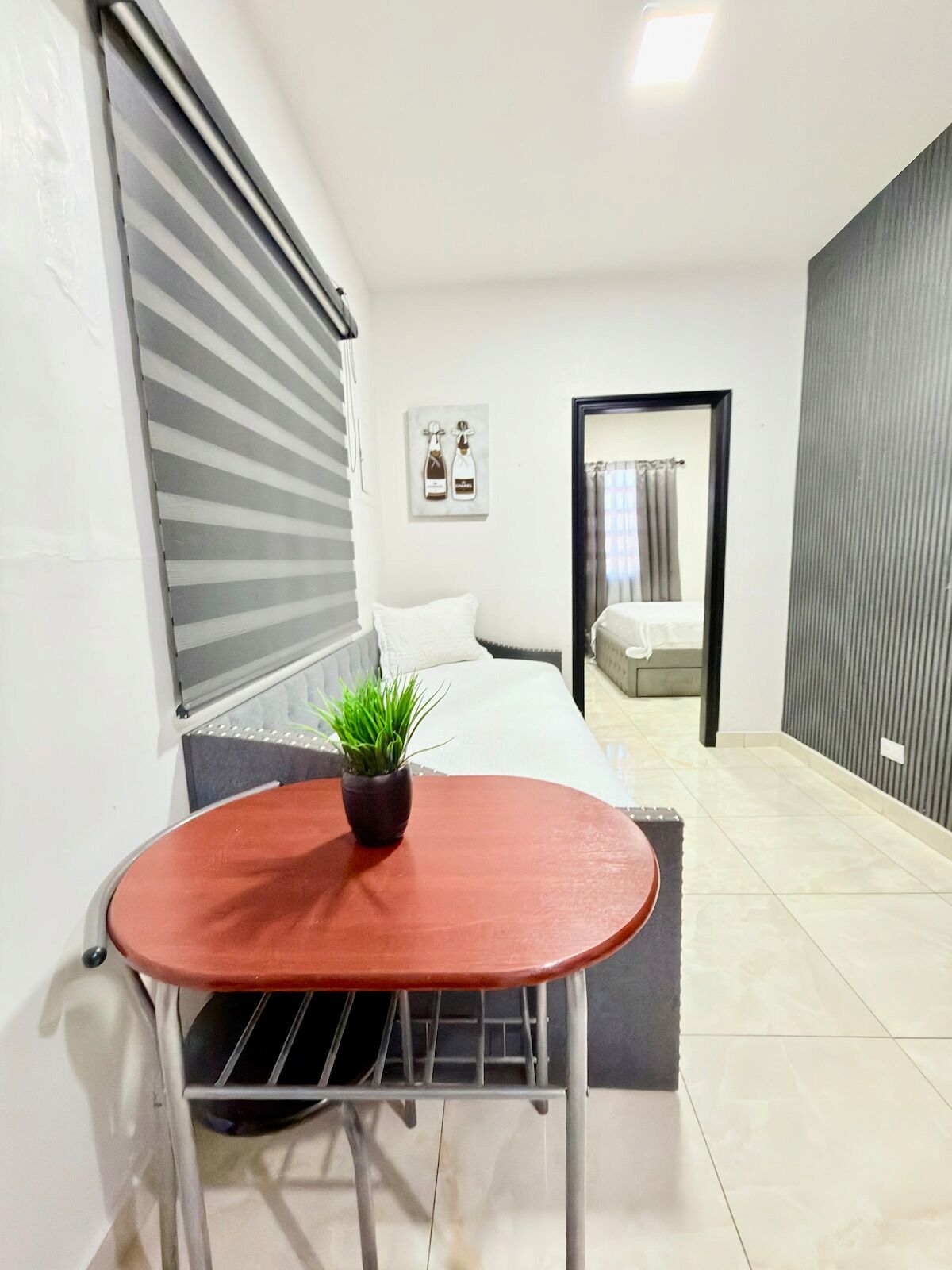 A compact room with a bed, a small table with a plant, and a window with blinds.