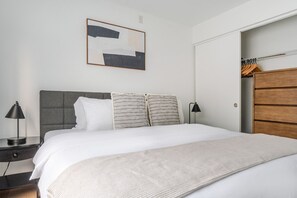 Uma cama bem arrumada, com cabeceira cinza, duas luminárias de apoio e um grande quadro abstrato na parede.