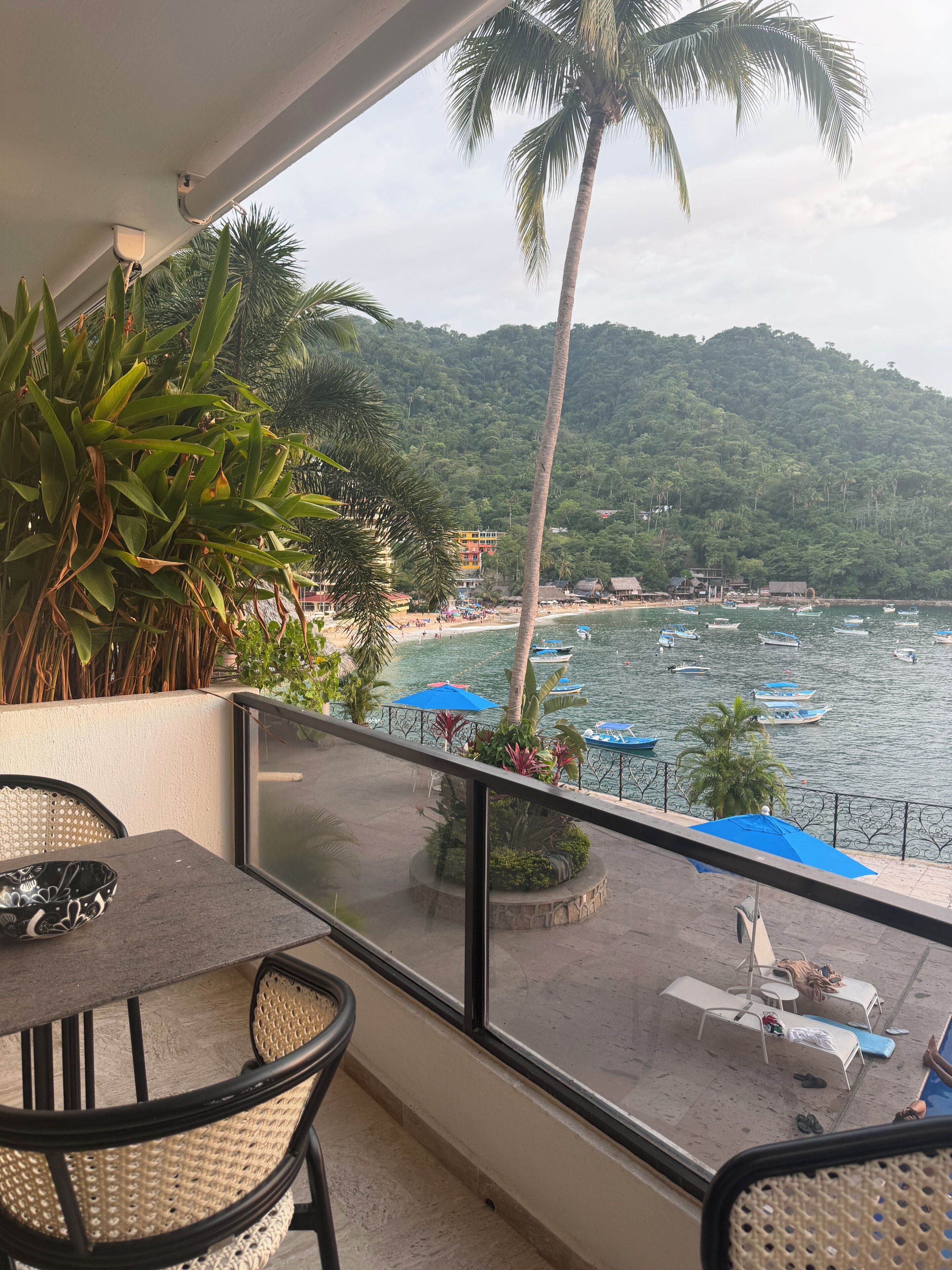 A balcony with a view of a beach and boats, featuring wicker furniture and potted plants.