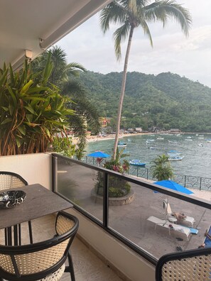 A balcony with a view of a beach and boats, featuring wicker furniture and potted plants. - Located right on the water! Modern 1-bedroom condo in tranquil Mismaloya  (Mismaloya)