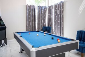 A pool table with balls, a blue chair, and a blue sofa in a room with a window and curtains. - Serene 5-bedrooms 3 bathrooms House with Pool in North Las Vegas (North Las Vegas)