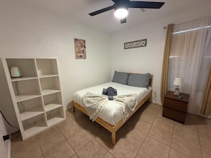 A bedroom with a bed, a nightstand, a shelf unit, and a ceiling fan.