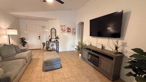 A living room with a sofa, ottoman, entertainment center, flat-screen TV, and potted plants. - Tutus House 🌞 Coldwater Springs, Avondale, AZ (Avondale)