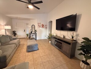 Ein Wohnzimmer mit Sofa, Hocker, Unterhaltungseinrichtung, Flachbildfernseher und Topfpflanzen.