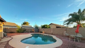 A swimming pool with a curved edge, surrounded by a pebble bed, with a patio area and outdoor furniture. - Tutus House 🌞 Coldwater Springs, Avondale, AZ (Avondale)
