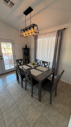 A dining area with a rectangular table, six chairs, a hanging light fixture, a potted plant, and a window with curtains. - Tutu's House in Coldwater Springs, Avondale, AZ (Avondale)
