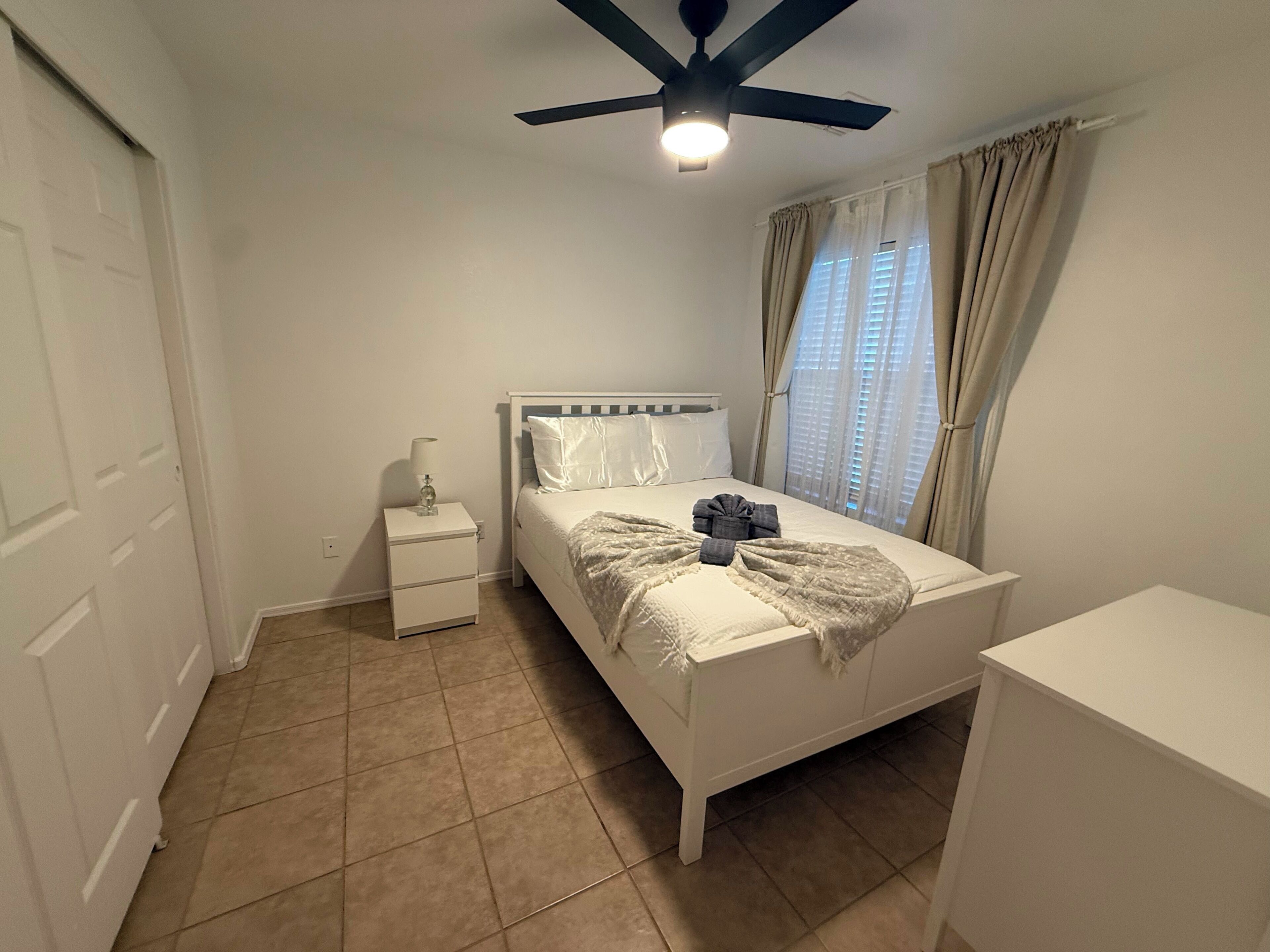 A bedroom with a bed, a nightstand, a ceiling fan, and a window with curtains.