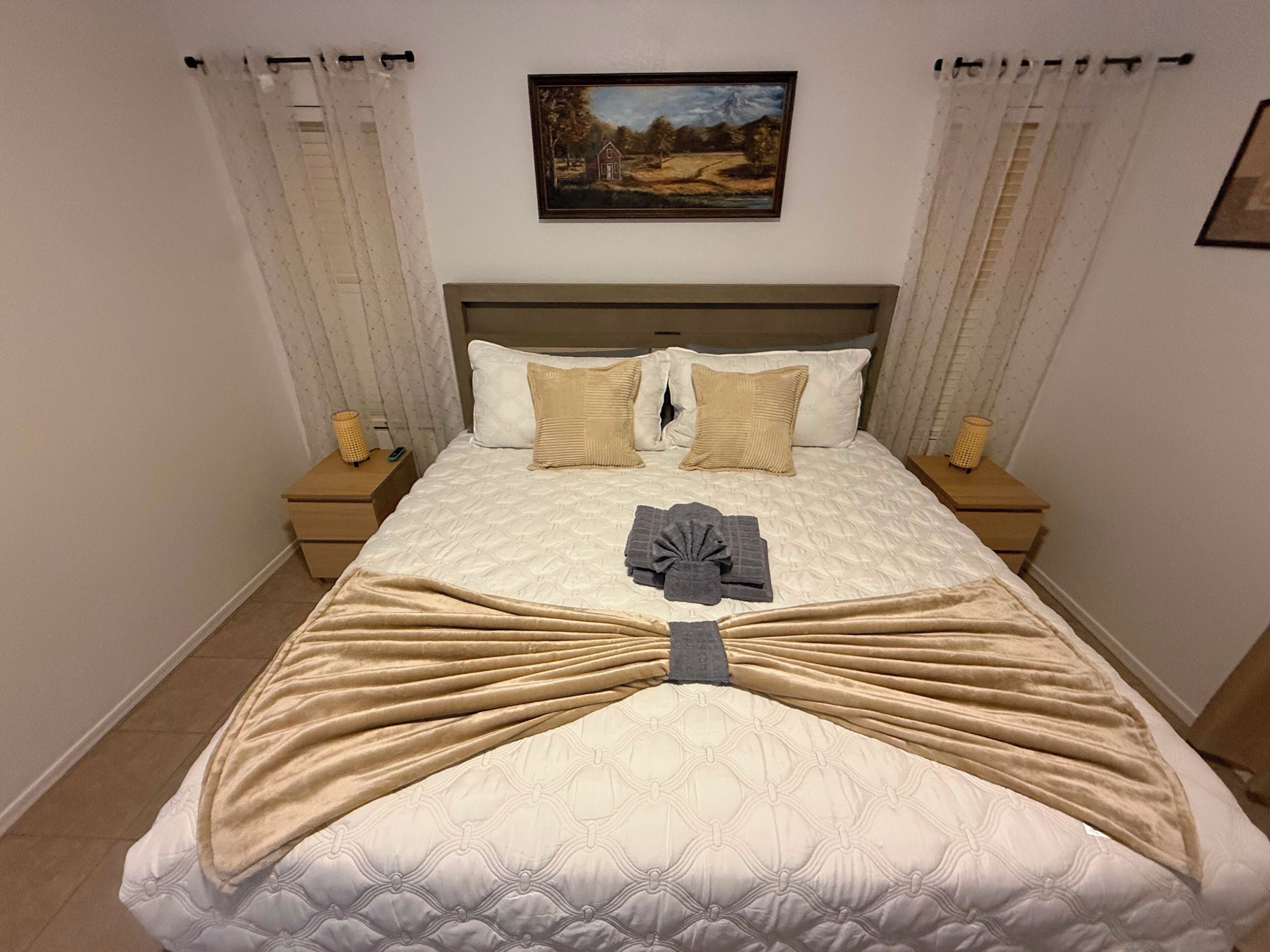 A neatly made bed with a quilt, pillows, and a folded towel. There is a painting on the wall above the bed.