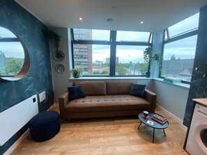 A modern living room with a brown sofa, a round mirror, a small round table with a book and a cup, and a washing machine. - Modern 1BR Apartment near MCR Stadiums (Stretford)