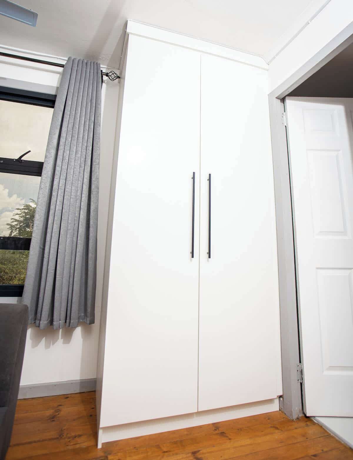 A white wardrobe with two doors and handles, a grey curtain, and a wooden floor.