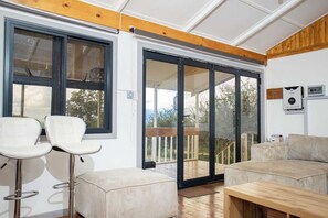 Una habitación con un gran ventanal corredero que da a una terraza exterior, un banco blanco acolchado y una mesa de madera.