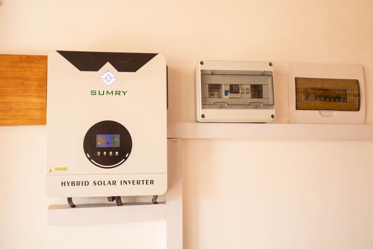 A hybrid solar inverter with a digital display, mounted on a wall above a white shelf.