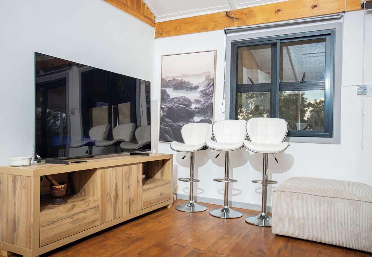 A modern living area with a wooden cabinet, a flat-screen TV, white bar stools, and a large window with a view.
