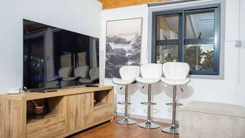 A modern living area with a wooden cabinet, a flat-screen TV, white bar stools, and a large window with a view.