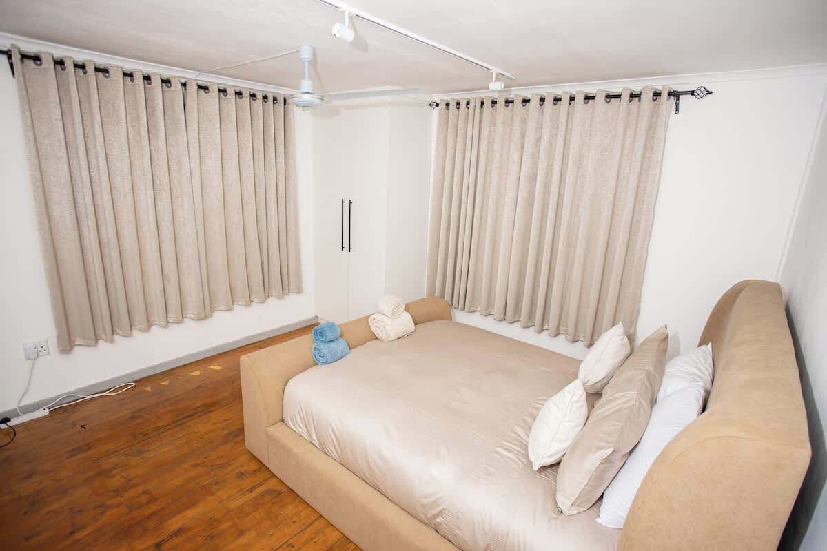 Une chambre avec un grand lit, des rideaux beiges, un ventilateur de plafond et un plancher en bois.