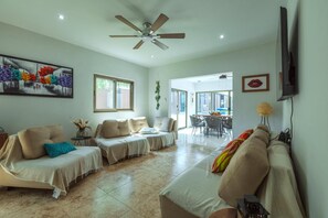 Un'ampia zona giorno con ventilatore a soffitto, TV a schermo piatto fissata al muro e sala da pranzo visibile tramite una porta aperta.