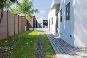 Un chemin menant à un bâtiment à l’extérieur blanc, avec une clôture marron et une zone bordée d’arbres.