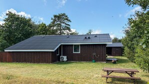 Ein hölzernes Bauernhaus mit geneigtem Dach, eine kleine angebaute Werkstatt, ein Picknicktisch und eine Rasenfläche.