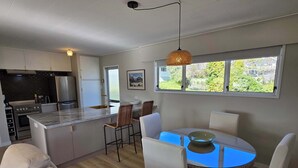 A modern kitchen with a central island, stainless steel appliances, and a dining area with white chairs and a glass table. - Comfy Haven in the Heart of Picton (Picton)