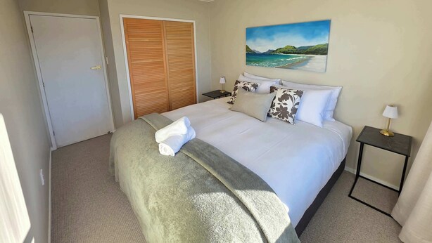 A hotel room with a bed, a painting of a beach, a wardrobe, and a nightstand with a lamp. - Comfy Haven in the Heart of Picton (Picton)