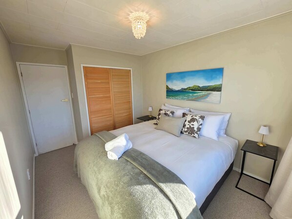 A hotel room with a bed, a painting of a beach, a wardrobe, and a nightstand with a lamp.