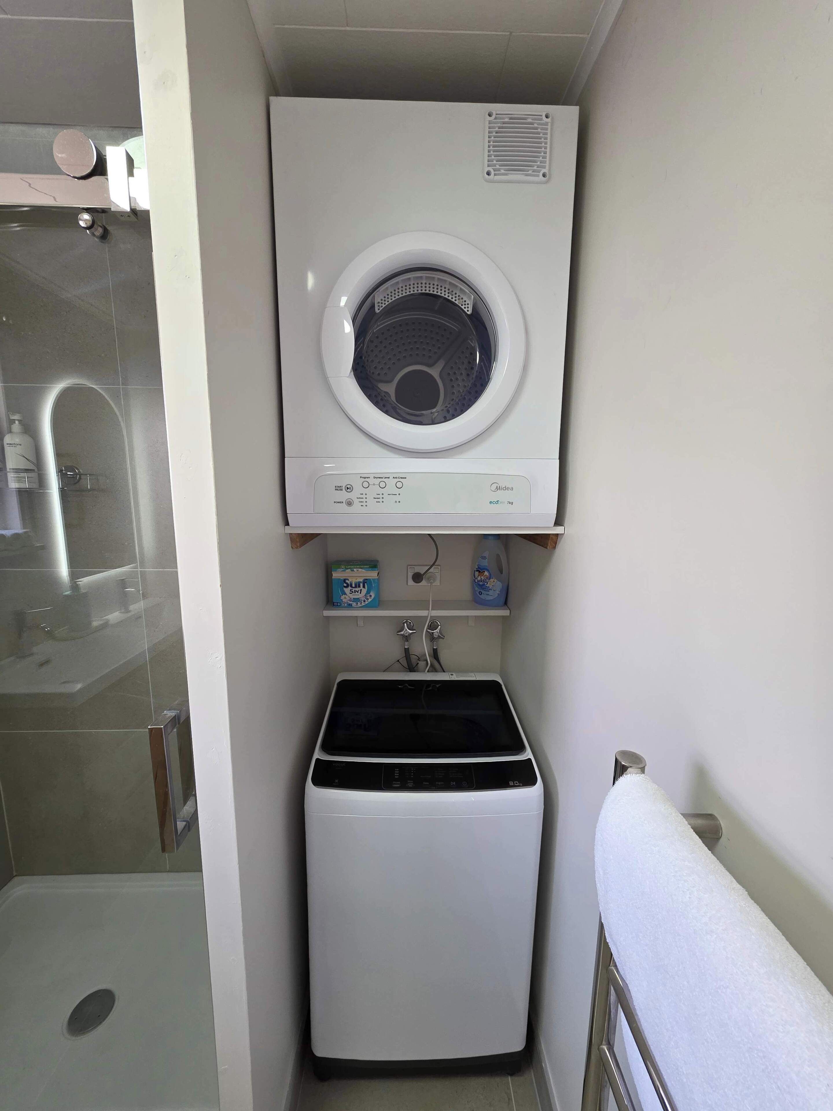 Een kleine wasruimte met een voorlader wasmachine, een droger en een klein plankje met schoonmaakspullen.