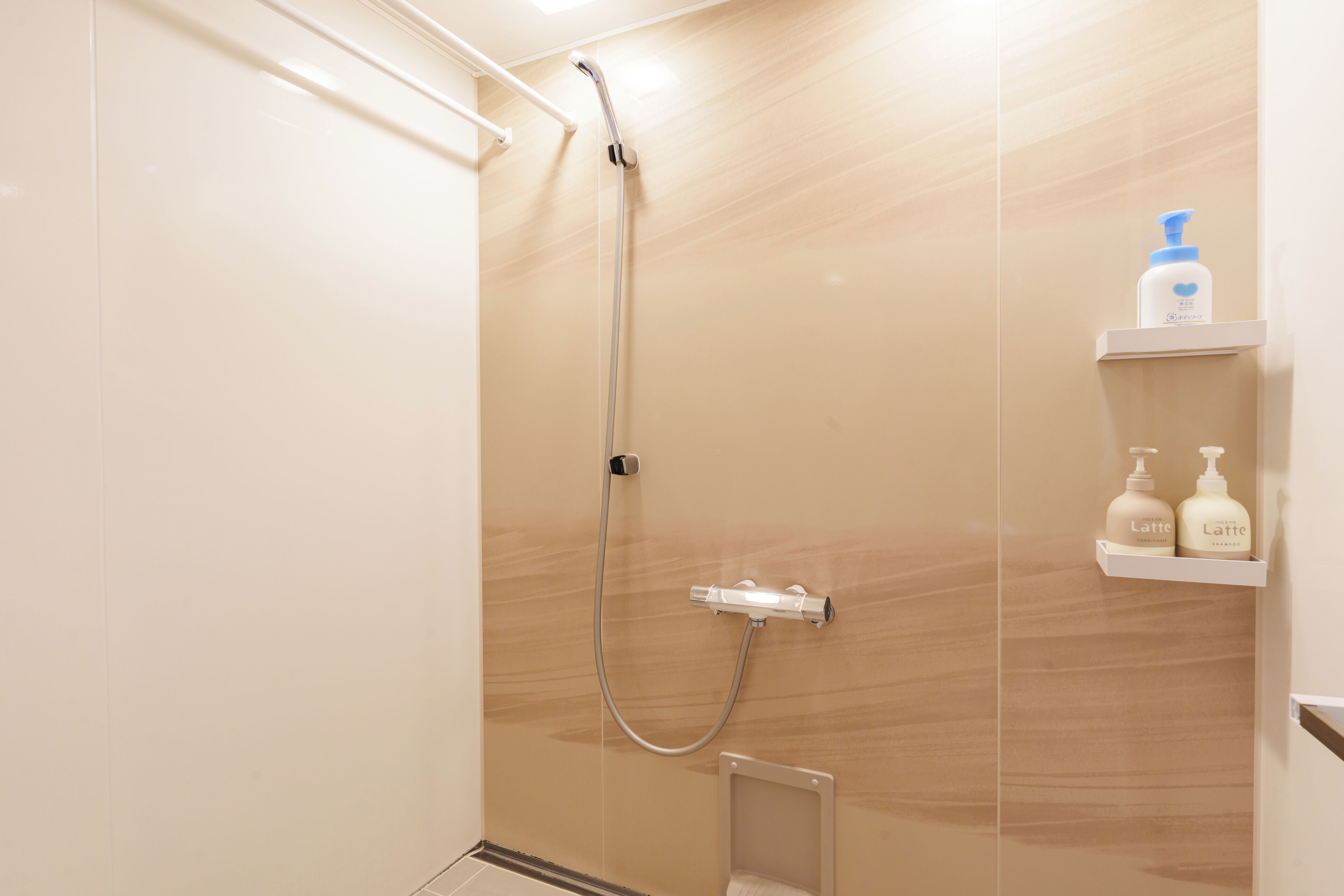 A shower area with a handheld showerhead and a wall-mounted control. There are two shelves with bottles of toiletries.