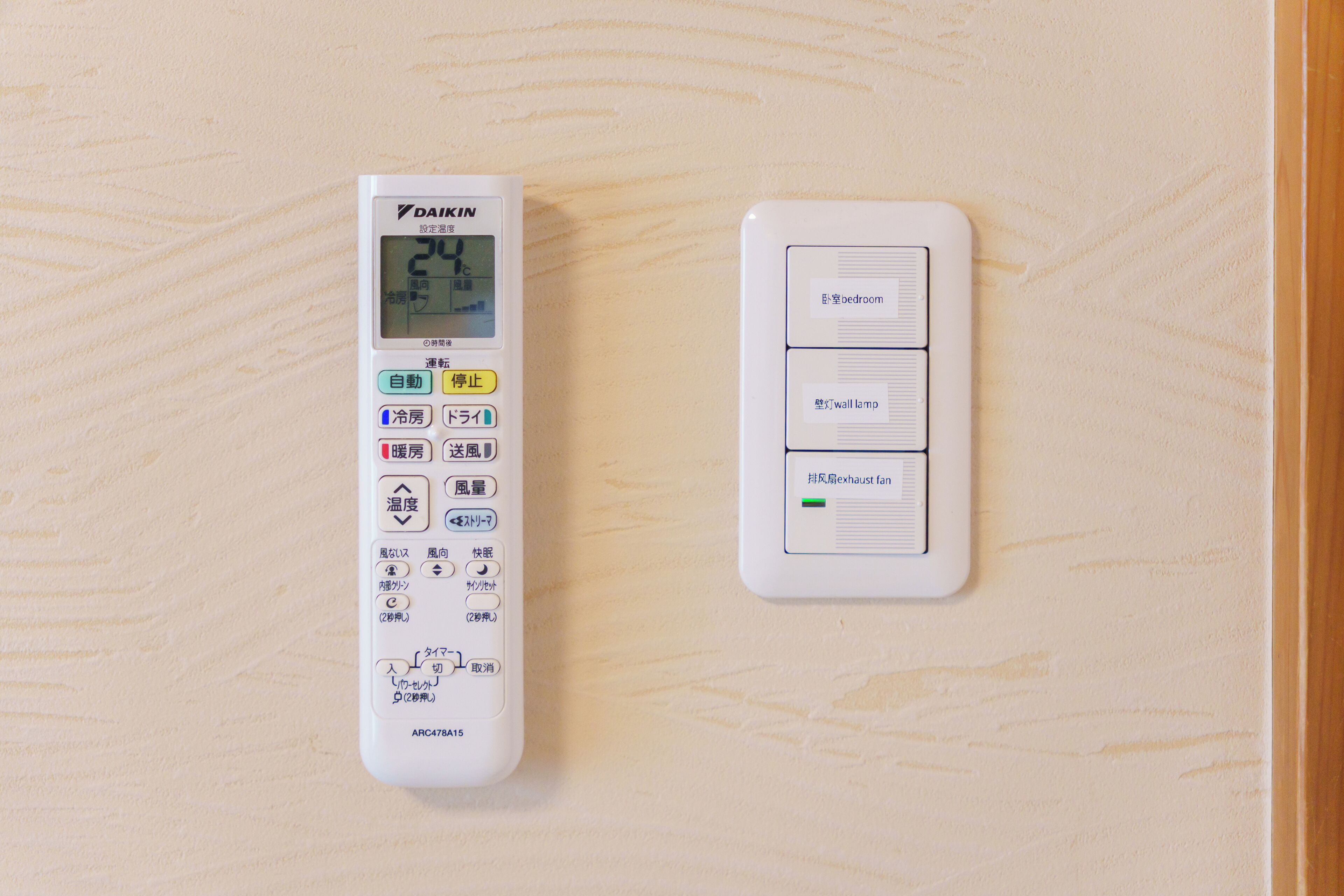 A wall-mounted thermostat and control panel with Japanese labels, indicating temperature settings and room functions.
