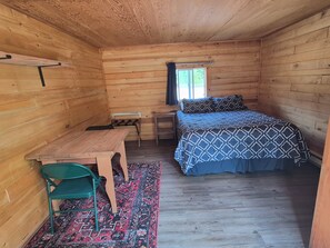 A small, wooden cabin room with a bed, a desk, a chair, and a window.
