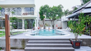 Une piscine avec des marches menant à l’eau, entourée d’un jardin planté de palmiers et d’un bâtiment doté d’un balcon.