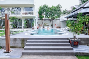 A swimming pool with steps leading up to it, surrounded by a garden with palm trees and a building with a balcony. - Gem's House (Mui Ne)