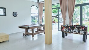 Une pièce équipée d’une table de billard, d’un baby-foot, d’un jeu de fléchettes et de grandes fenêtres avec des rideaux.