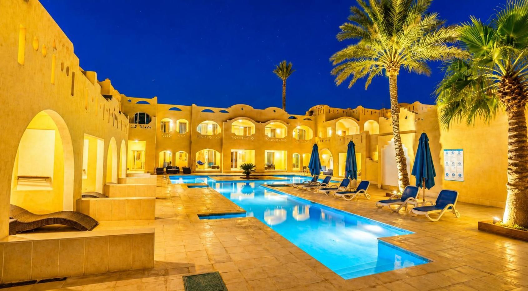 A swimming pool with lounge chairs and palm trees, surrounded by a building with arches and warm lighting.